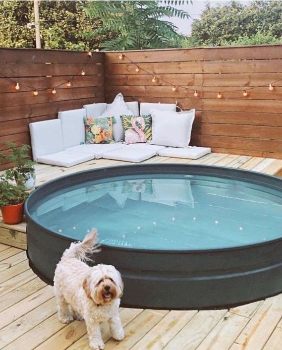 A cool outdoor space with a wooden deck, walls with lights, a stock tank pool, some pillows, cushions and potted plants.