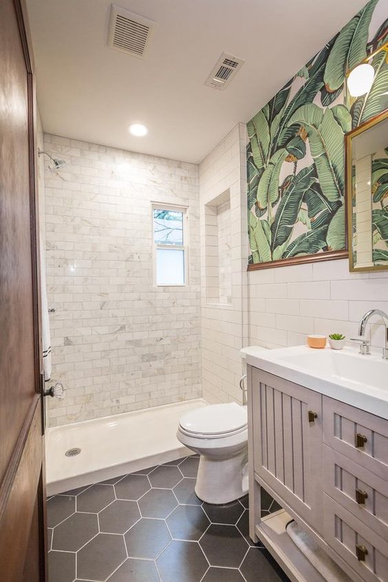 A cool small bathroom with white marble and black hexagon tiles, a pale pink vanity, tropical print wallpaper and a mirror in a gold frame.