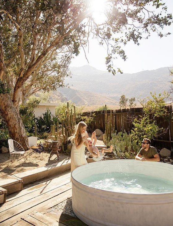 A cozy desert backyard with growing cacti and a tree, a wooden deck, a white stock tank pool is a lovely space to enjoy.