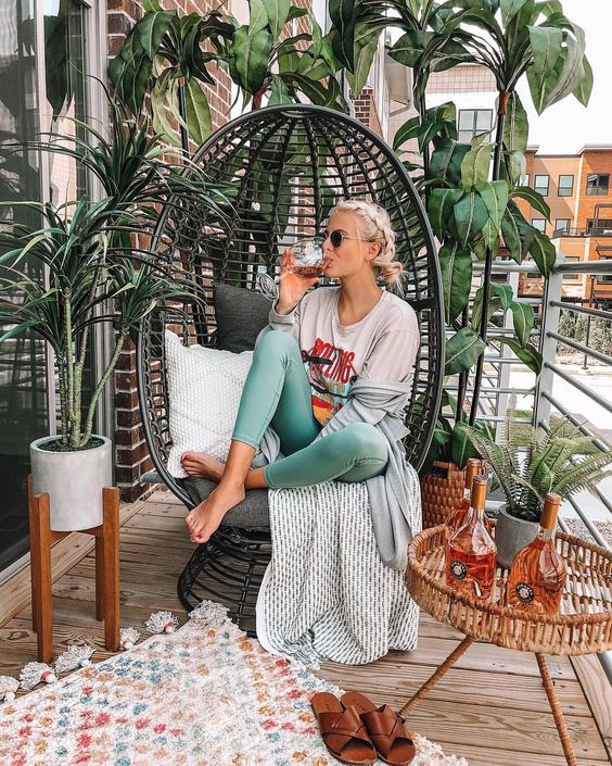 A cute small balcony with a large rattan egg chair and a rattan table, some statement plants and cozy textiles.