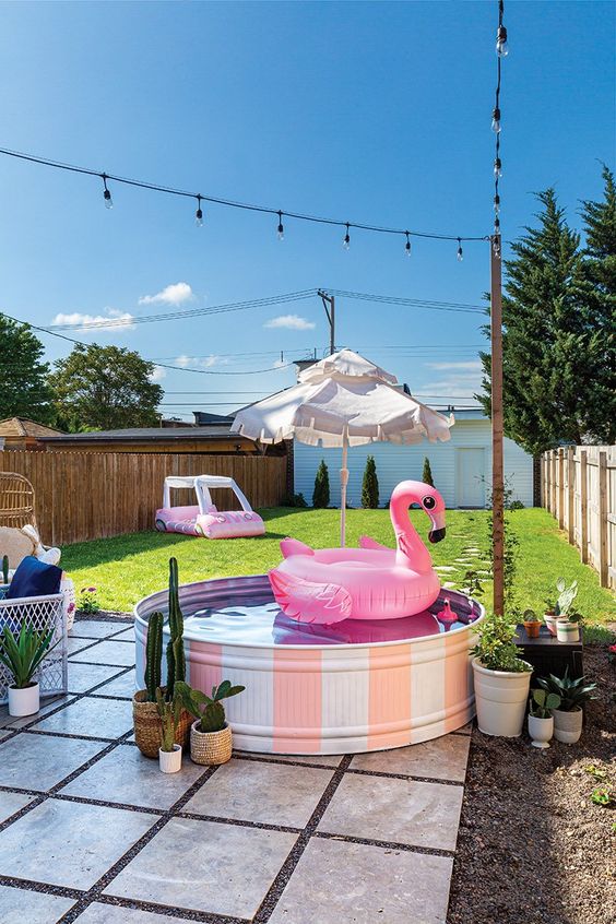 A green lawn, a tiled deck with a stock tank pool and potted plants, a rattan chair and some string lights.
