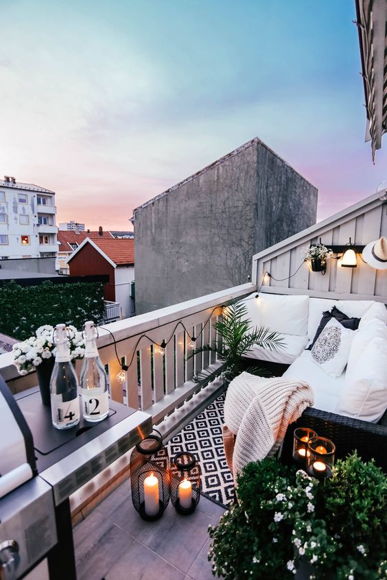 A monochromatic small balcony with black wicker furniture, candle lanterns, potted greenery and blooms and a grill.