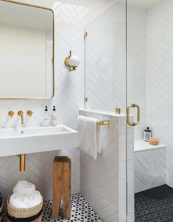 A refined bathroom with a dark floor, a shower space with a pony wall, white herringbone tiles and gold touches here and there.