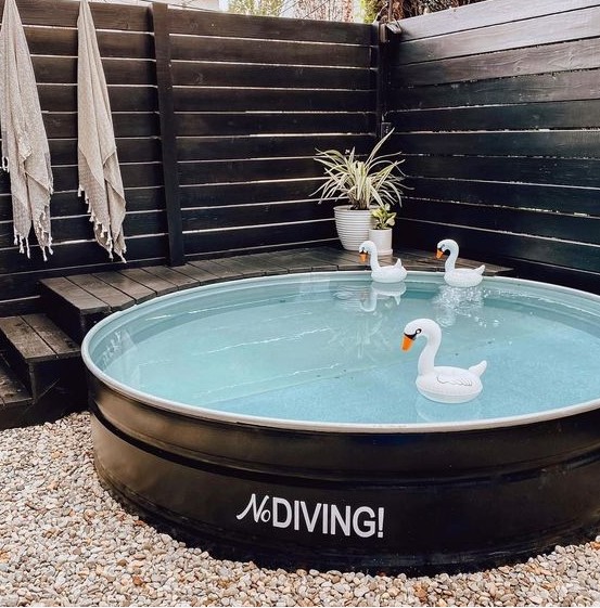 A small and cute pool nook with a black stock tank pool with floats, a ladder and a deck and planked screens around the pool.