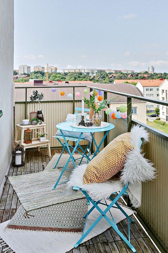 A small and cute space with blue metal furniture, layered rugs, printed pillows, lights, candle lanterns and greenery.