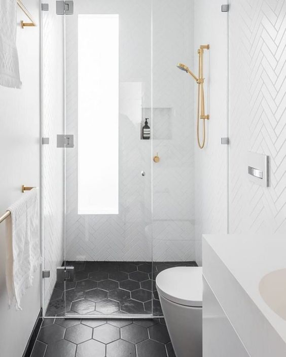 A small bathroom with white herringbone tiles and black hex ones, with a frosted glass window and gold fixtures.