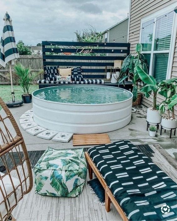 A tropical outdoor space with a black bench and pillows, a white stock tank pool, tiles, a lounger and tropical plants.