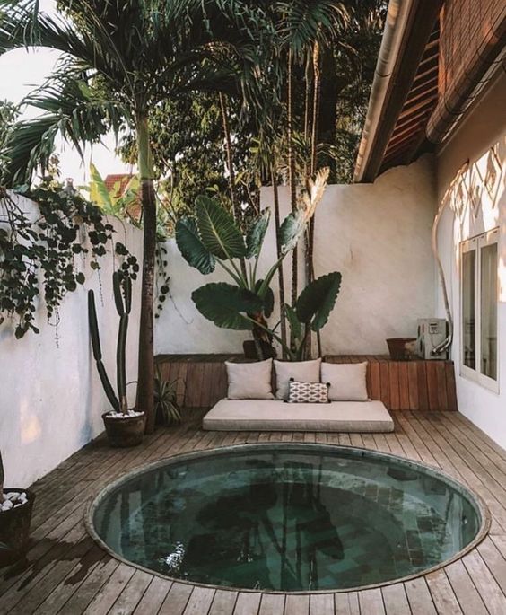 A tropical outdoor space with an in-ground stock tank pool, a wooden deck, a cushion, potted plants and trees.