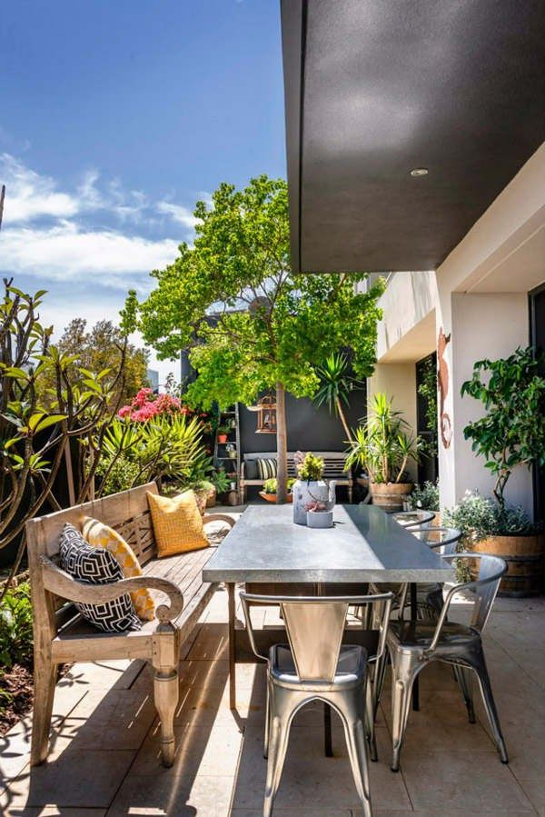 A tropical terrace with lots of greenery and trees, a vintage carved wooden bench and metal furniture, bright pillows.