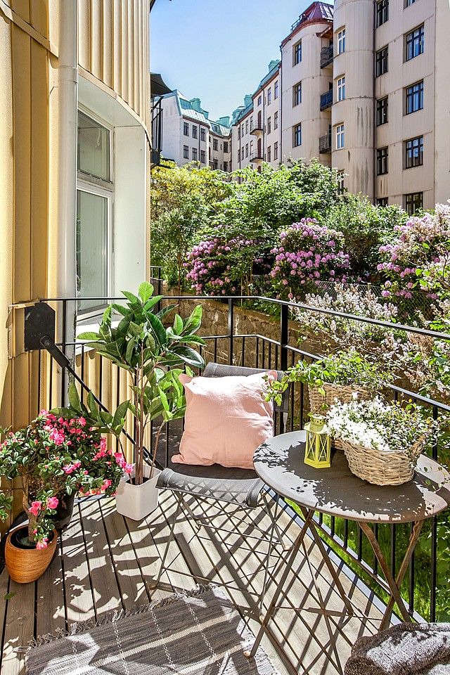 A tiny balcony with lots of potted greenery and blooms and some metal furniture plus some pillows.