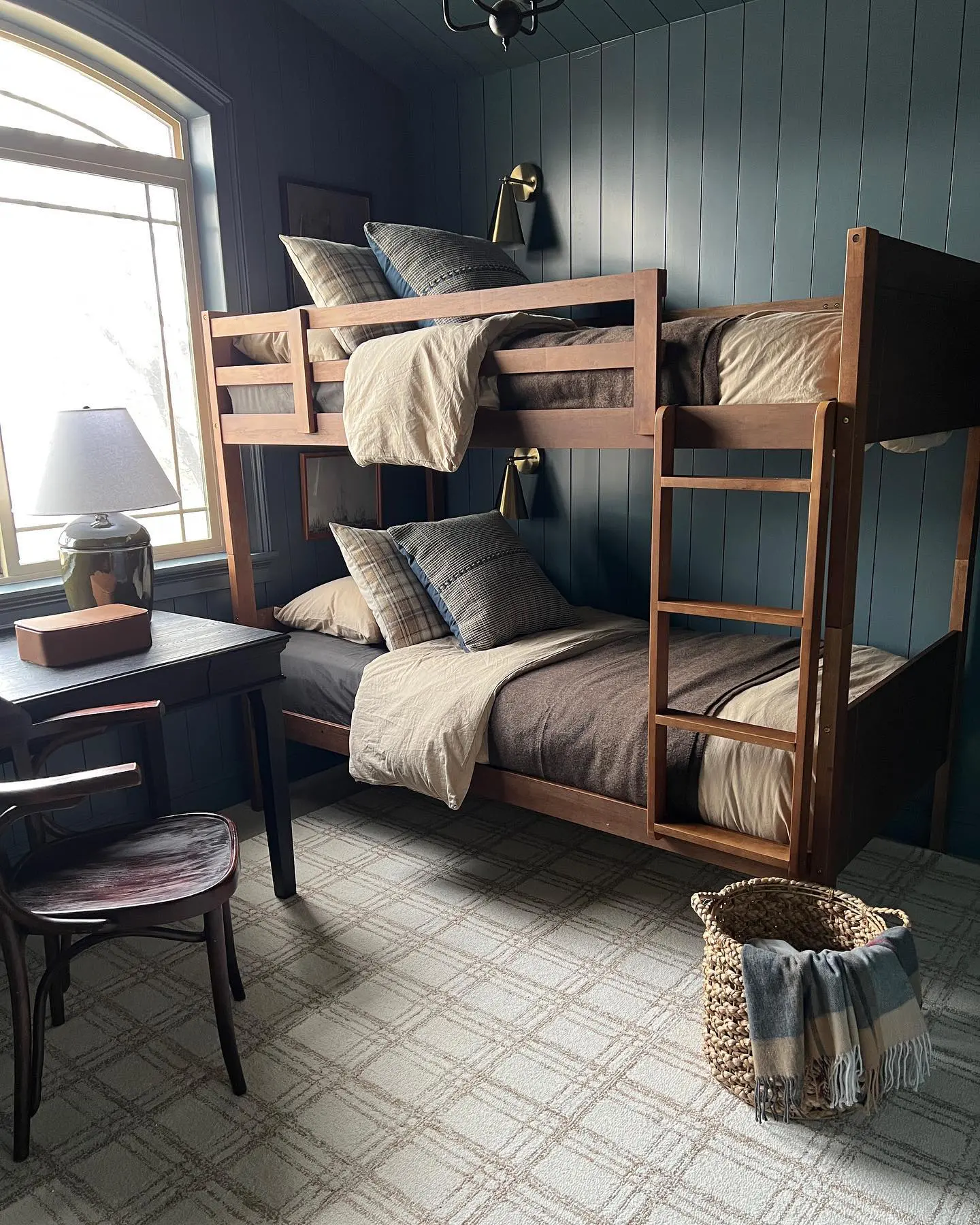This beautifully designed shared bedroom exudes a rich, moody country charm. The dark green walls create a cozy atmosphere, while natural light from the large window keeps the space bright. The wooden bunk beds paired with neutral bedding and checkered accents enhance the rustic appeal. 

(@house.on.oak.lane).