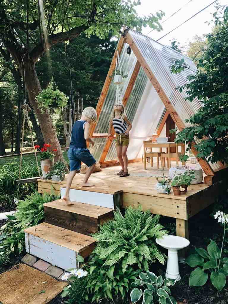 This kids' house is composed of pallets and glass, there's a small set of furniture and lots of greenery around.